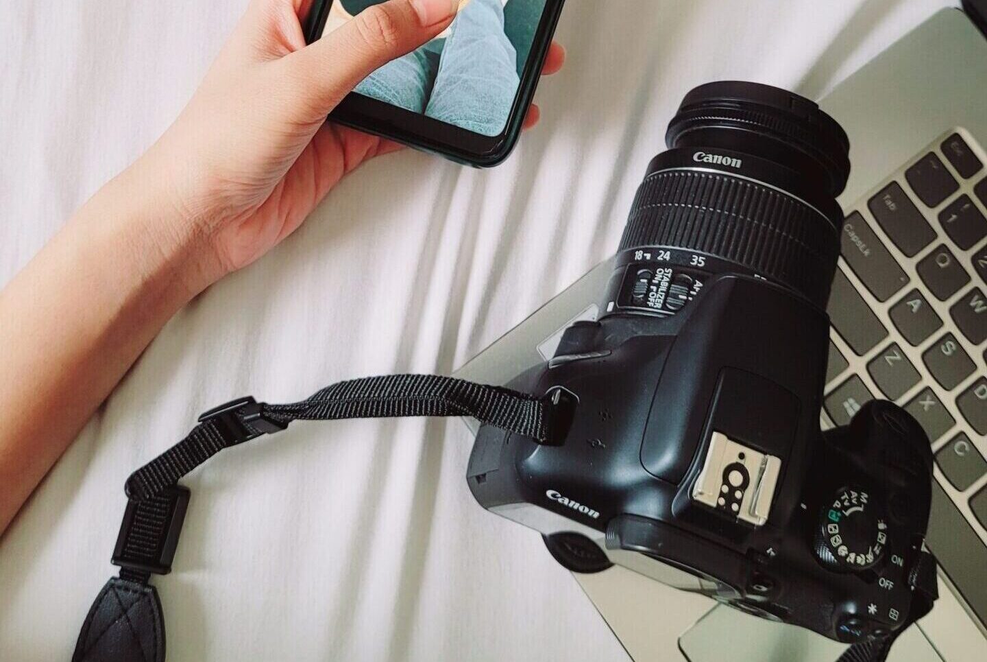 A hand holding a smartphone above a DSLR camera and laptop on a white surface, showcasing modern technology and photography.